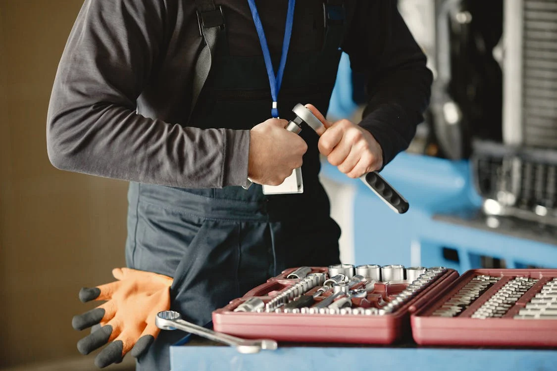 a worker dressed in dark overalls and a long-sleeve shirt, holding a ratchet wrench while working on something out of frame. A red toolset containing various sockets and tools is open on a blue surface in the foreground. An orange pair of work gloves rests beside the toolset