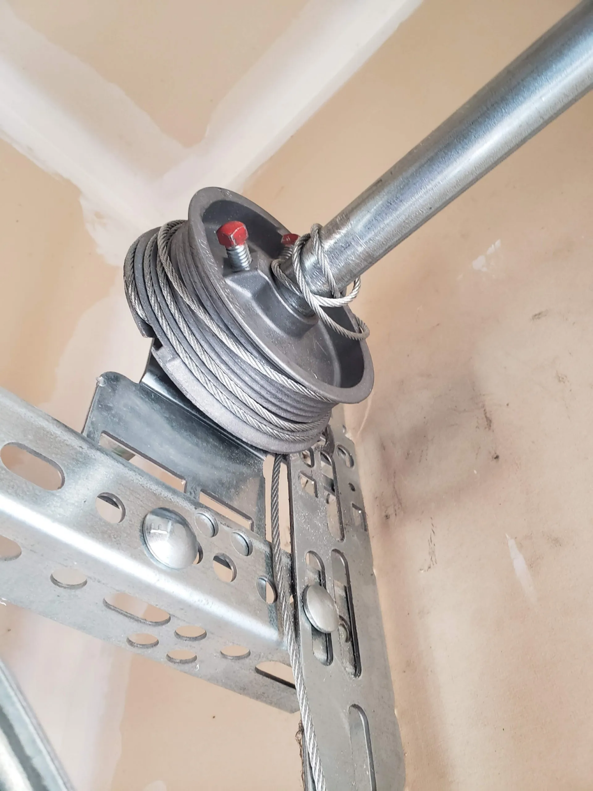 a close-up view of a metal pulley system, typically used in mechanical or garage door setups. The system includes a coiled steel cable wound around a pulley, secured with a red bolt or pin. The pulley is attached to a metal bracket with a series of slots and bolts for adjustment and support