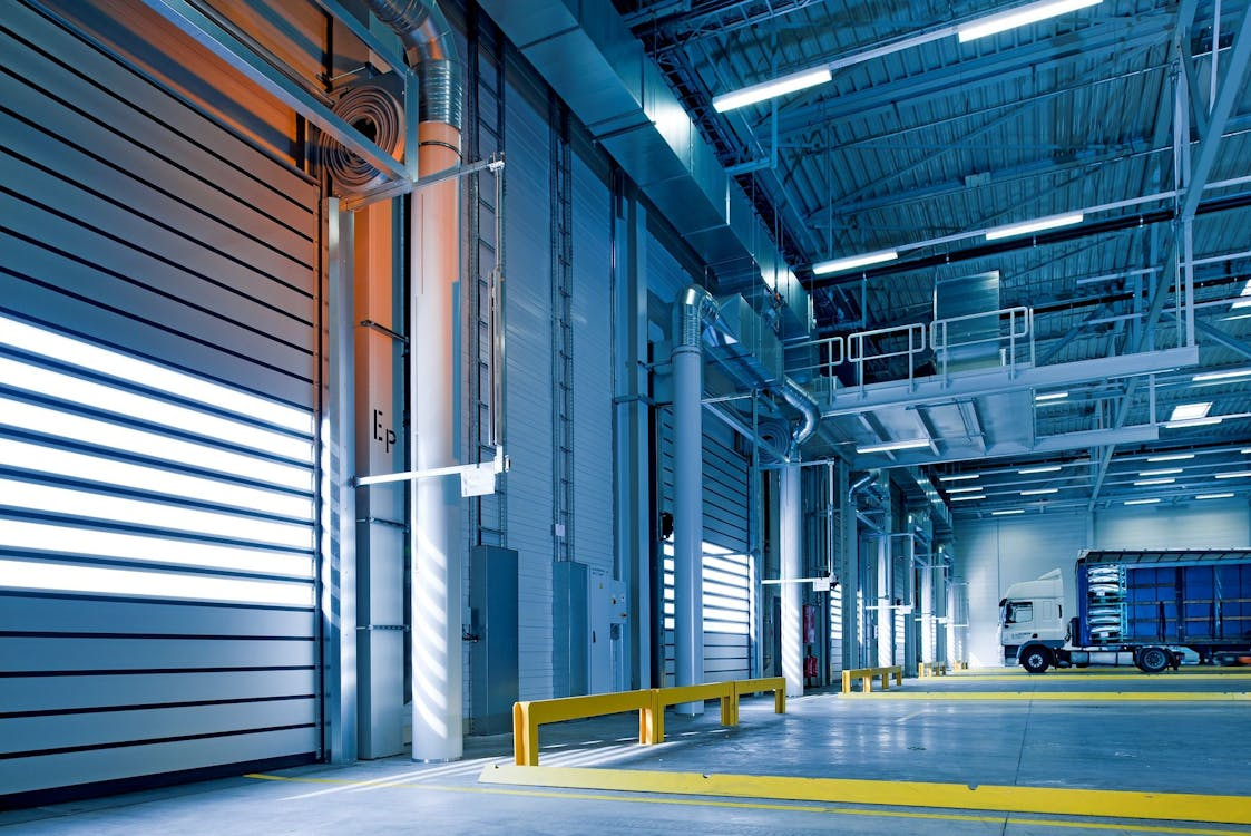 a modern, well-lit industrial or warehouse interior. The space features high ceilings with exposed steel beams and bright fluorescent lighting. On the left, large sectional doors with horizontal panels are visible, illuminated by external light. The space is organized, with yellow safety barriers lining the pathways, ensuring clear separation between vehicle and pedestrian zones