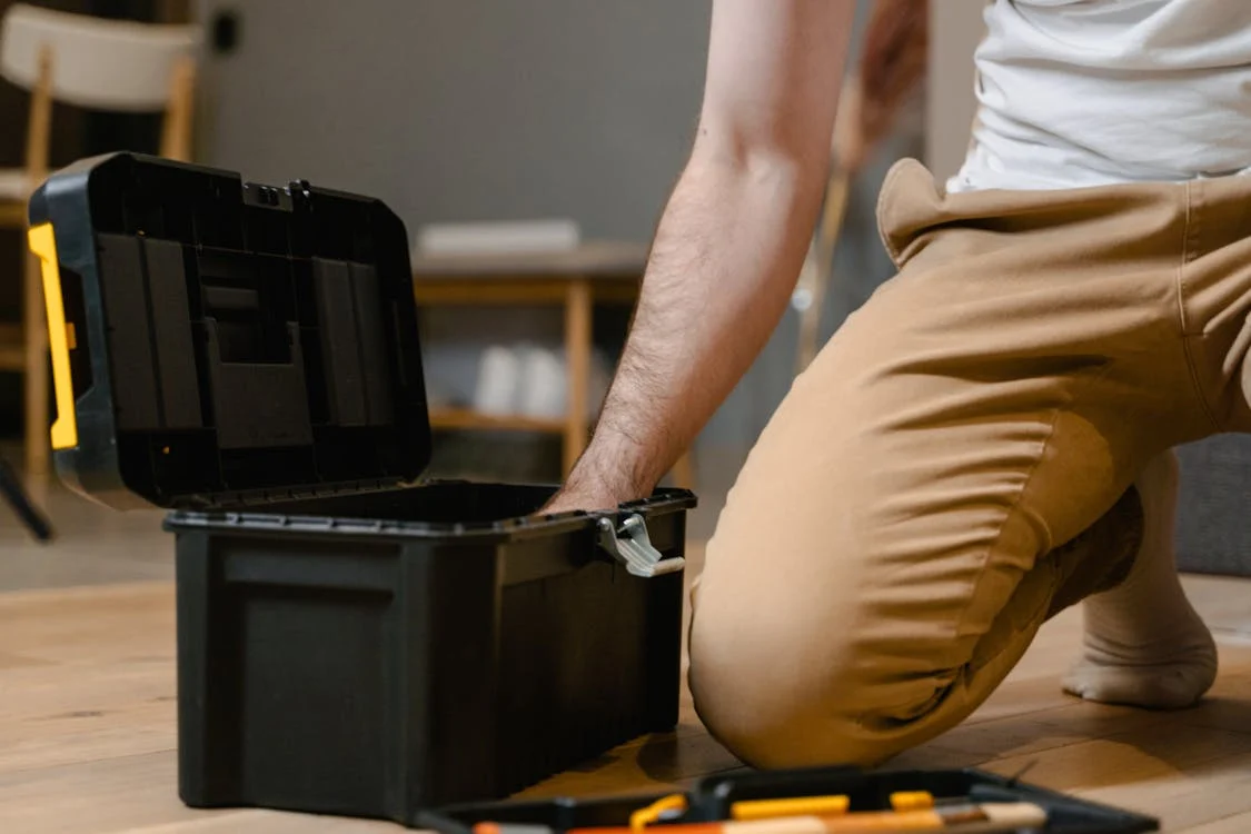 a person kneeling on a wooden floor while reaching into an open black toolbox with yellow accents. The scene suggests the individual is in the middle of a repair or assembly task. The environment appears to be indoors, likely a home setting, with blurred furniture visible in the background, including a chair and shelving